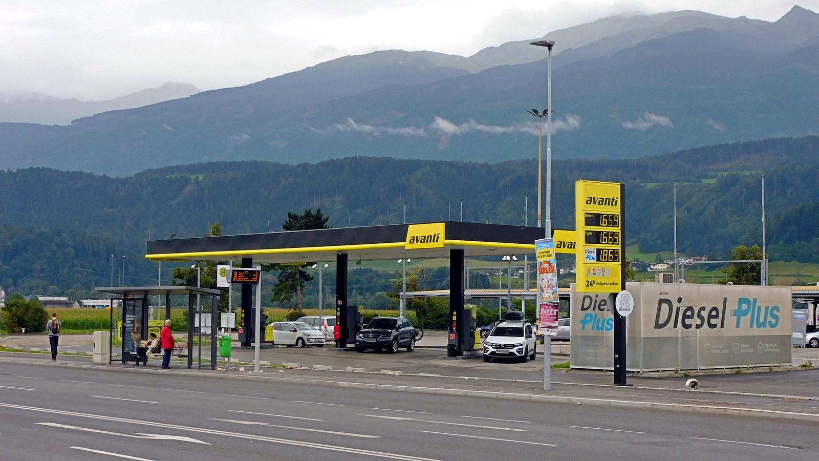 Tankstelle mit schwarz-gelbem Dach und Preistafel in Neu-Rum bei Innsbruck, dahinter bewaldete Berge unter Wolken